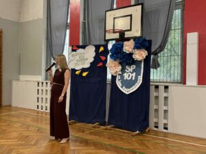 Na zdjęciu znajduje się Pani Dyrektor przemawiająca do uczniów zgromadzonych na uroczystym rozpoczęciu roku szkolnego 2026/2027. Trzyma w ręku mikrofon i uśmiecha się. Ubrana jest w elegancki bordowy kombinezon. Stoi na środku sali gimnastycznej. Za nią znajduje się dekoracja z kwiatów wykonanych z bibuły oraz napis "Rozpoczęcie roku szkolnego 2026/2027"
