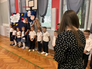 Na zdjęciu znajduje się grupa dziewięciu uczniów pozujących do wspólnego zdjęcia wraz ze swoimi opiekunami. Stoją na tle dekoracyjnej ścianki przygotowanej z okazji rozpoczęcia roku szkolnego. Uczniowie ubrani są w stroje galowe i uśmiechają się do zdjęcia