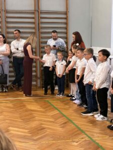 Na zdjęciu znajduje się grupa siedmiu uczniów ubranych w stroje galowe. Pani Dyrektor wita się z nimi, podając im rękę. W tle widoczni są rodzice obserwujący dzieci